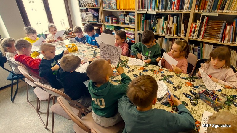 W czytelni biblioteki przy długim stole nakrytym roślinnym obrusem siedzi kilkanaście dzieci. Na blacie leżą okrągłe, czarno‑białe karty z&nbsp;roślinnymi wzorami i&nbsp;motylami, kredki oraz nożyczki. Dzieci kolorują i&nbsp;wycinają elementy do kompozycji „wiosenna łąka”. W&nbsp;tle widać regały z&nbsp;książkami oraz okna z&nbsp;widokiem na ulicę.