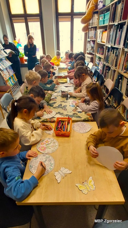 Przy dwóch złączonych stołach siedzi liczna grupa dzieci, każde pracuje nad własną kartą: ilustracją w&nbsp;okręgu z&nbsp;roślinnymi motywami i/lub papierowym motylem do przyklejenia na „wiosenną łąkę”. Na środku leżą koszyki z&nbsp;kredkami i&nbsp;nożyczkami. W&nbsp;tle – regały pełne książek oraz okna doświetlające przestrzeń. 