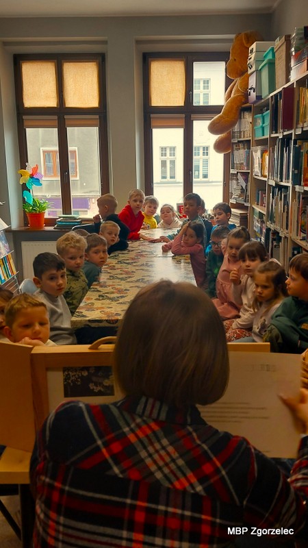 Na pierwszym planie widać drewnianą skrzynkę teatru kamishibai ustawioną na brzegu stołu. Po obu jego stronach siedzą dzieci, zwrócone w&nbsp;kierunku prezentacji. W&nbsp;tle znajdują się wysokie regały z&nbsp;książkami, okna z&nbsp;zasłoniętymi roletami oraz duża maskotka na półce. Zajęcia odbywają się w&nbsp;ramach marcowego cyklu o&nbsp;wiośnie.