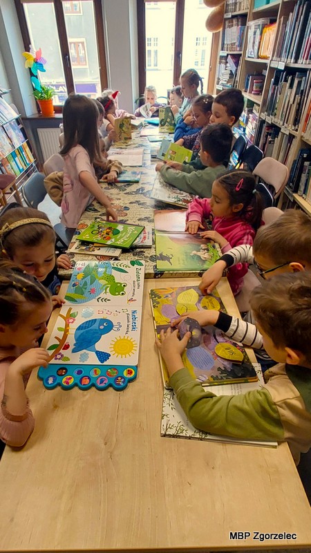 W bibliotece ustawiono dwa długie stoły, przy których siedzi grupa dzieci trzymających w&nbsp;dłoniach książki z&nbsp;kolorowymi ilustracjami. Widoczne są książki o&nbsp;zwierzętach, roślinach i&nbsp;zabawnych bohaterach. Za dziećmi stoją regały z&nbsp;literaturą dziecięcą, a&nbsp;obok dużych okien znajduje się dekoracja w&nbsp;kształcie kwiatka.