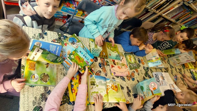 Dzieci siedzą blisko siebie po obu stronach stołu nakrytego obrusem w&nbsp;motyle. Na blacie rozłożone są liczne książki — część otwarta na dużych ilustracjach przedstawiających rośliny, zwierzęta i&nbsp;scenki z&nbsp;książek edukacyjnych. W&nbsp;tle widać biblioteczne regały pełne książek.