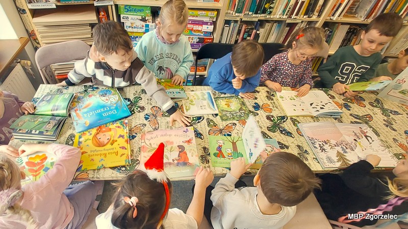 Przy długim stole w&nbsp;czytelni siedzi kilkanaście dzieci, każde ma przed sobą inną książkę obrazkową. Na stole leżą publikacje o&nbsp;wiośnie, roślinach, zwierzętach i&nbsp;codzienności przedszkolaków. Dzieci kartkują strony, porównują ilustracje i&nbsp;pokazują sobie znalezione obrazki. W&nbsp;tle widać regały z&nbsp;książkami.