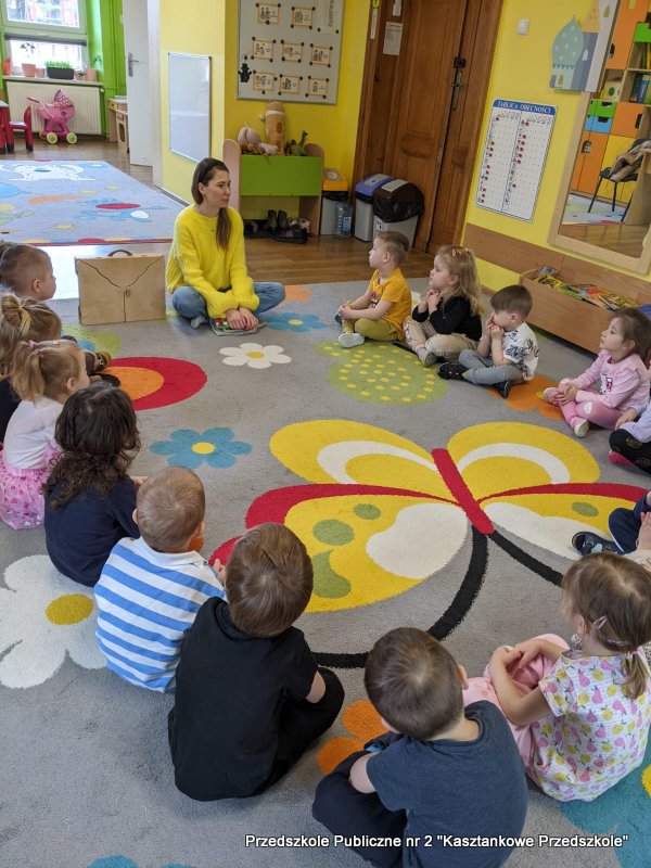 W przestronnej sali przedszkolnej dzieci siedzą wokół bibliotekarki, tworząc krąg na dywanie z&nbsp;dużym wzorem motyla i&nbsp;kwiatami. W&nbsp;tle widać tablicę, zabawki, małe stoły i&nbsp;krzesła. Po jednej stronie znajdują się otwarte półki z&nbsp;materiałami edukacyjnymi i&nbsp;pojemnikami.
