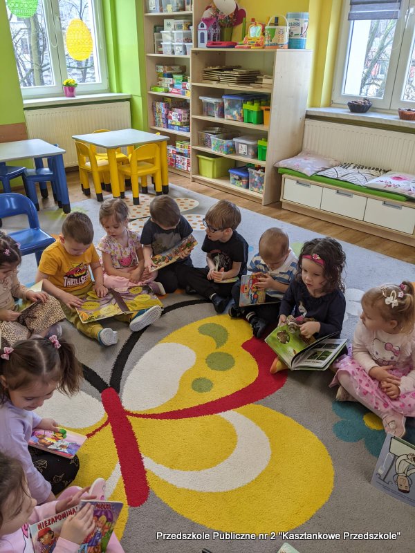 Dzieci siedzą na kolorowym dywanie w&nbsp;sali przedszkola, każde trzyma własną książkę z&nbsp;ilustracjami. Obok nich stoją małe kolorowe krzesełka, a&nbsp;z tyłu widoczny jest niski stół oraz półki z&nbsp;zabawkami i&nbsp;materiałami edukacyjnymi. Książki mają duże, kolorowe obrazki, które dzieci pokazują sobie nawzajem.