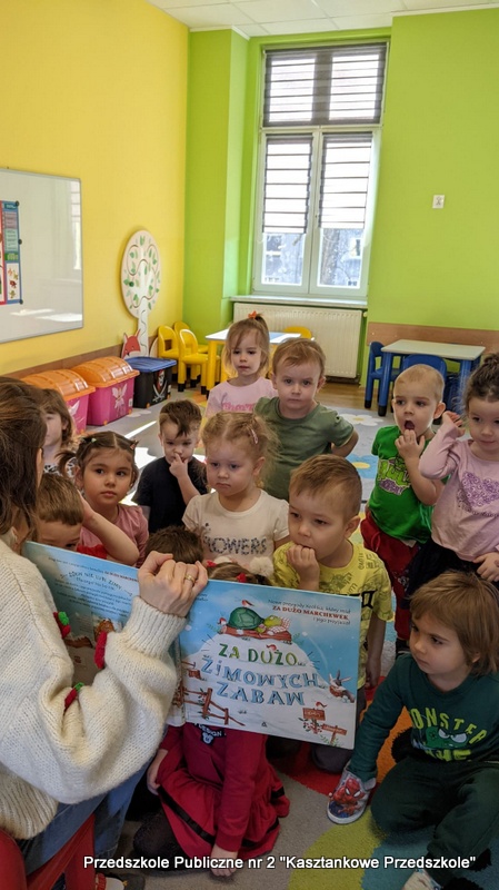 W sali przedszkolnej dzieci siedzą blisko bibliotekarki, która trzyma otwartą książkę „Za dużo zimowych zabaw”. Pomieszczenie jest jasne, z&nbsp;zielonymi i&nbsp;żółtymi ścianami, wyposażone w&nbsp;małe stoliki, krzesełka i&nbsp;zabawki. Dzieci skupiają się na oglądaniu prezentowanej książki.