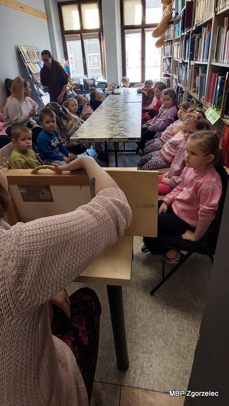 W czytelni biblioteki dzieci siedzą wzdłuż stołu i&nbsp;patrzą w&nbsp;stronę drewnianej skrzynki kamishibai, ustawionej na krótszym boku stołu. Po obu stronach znajdują się regały z&nbsp;książkami, a&nbsp;za dziećmi duże okna doświetlają pomieszczenie. Na pierwszym planie widać bibliotekarkę zajęcia w&nbsp;trakcie prezentacji teatrzyku obrazkowego.