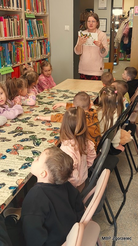 Dzieci siedzą przy stole nakrytym obrusem w&nbsp;motyle i&nbsp;zwracają się w&nbsp;stronę bibliotekarki prowadzącej, stojącej przy wejściu do pomieszczenia. Bibliotekarka pokazuje ilustrację motyla. W&nbsp;tle znajdują się regały z&nbsp;książkami oraz korytarz prowadzący do dalszej części biblioteki.