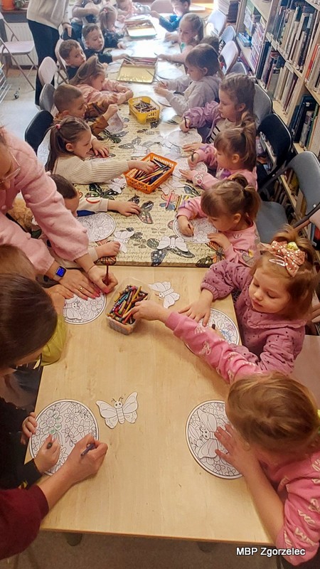 Przy dwóch złączonych stołach w&nbsp;czytelni biblioteki siedzi grupa dzieci, które kolorują okrągłe ilustracje z&nbsp;roślinnymi wzorami oraz papierowe motyle. Na stołach leżą kredki, koszyki z&nbsp;przyborami i&nbsp;przygotowane szablony prac. W&nbsp;tle stoją wysokie regały wypełnione książkami.