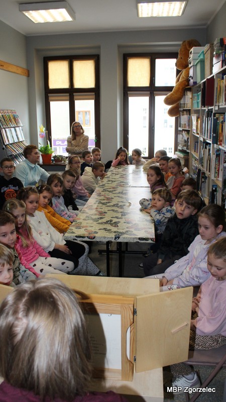 Dzieci siedzą przy stole w&nbsp;bibliotece i&nbsp;oglądają teatrzyk kamishibai ustawiony przed nimi