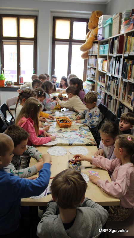 Grupa dzieci siedzi przy stole w&nbsp;bibliotece i&nbsp;koloruje okrągłe ilustracje, korzystając z&nbsp;kredek i&nbsp;materiałów plastycznych