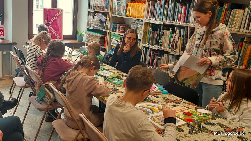Dzieci i&nbsp;dorośli pracują przy stole w&nbsp;bibliotece, kolorując ilustracje i&nbsp;wykonując zadania plastyczne związane z&nbsp;tematyką wiosenną. W&nbsp;tle widać regały z&nbsp;książkami.