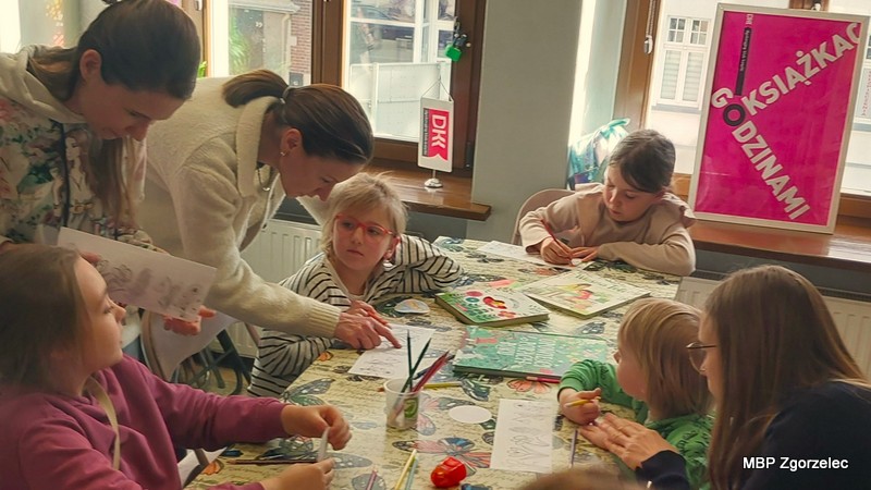 Dzieci siedzą przy stole, rysując i wyklejając papierowe elementy o motywach roślinnych, a trzy osoby dorosłe pomagają im w pracy twórczej. Na stole rozłożone są kredki, papier i materiały plastyczne.