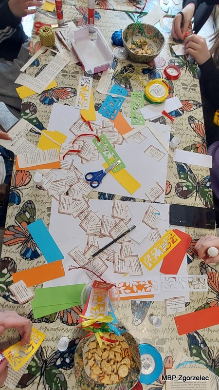 Widok z&nbsp;góry na stół przykryty obrusem, na którym rozłożone są papierowe zakładki, kolorowe kartony, nożyczki, klej, szablony i&nbsp;fragmenty tekstów z&nbsp;książek. Kilka osób pracuje przy wspólnym stole, wykonując zakładki do książek dla czytelników Miejskiej Biblioteki Publicznej w&nbsp;Zgorzelcu.