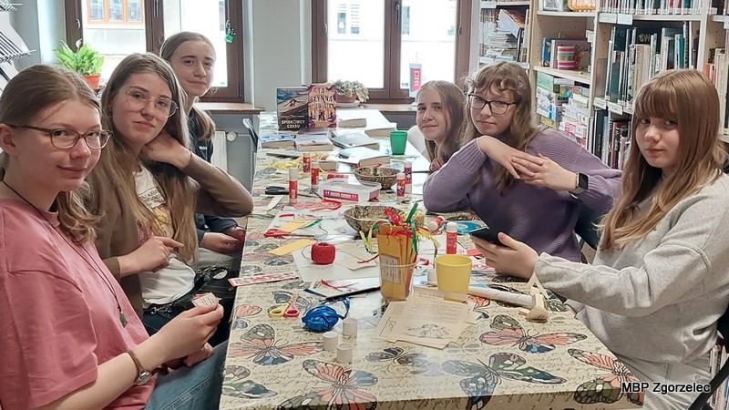 Uczestniczki siedzą przy długim stole w przestrzeni bibliotecznej, pomiędzy regałami z książkami. Na stole znajdują się materiały plastyczne, papier, zakładki i przybory do prac ręcznych. Zdjęcie przedstawia spotkanie Młodzieżowego Klubu „Książkowi Wędrowcy”, podczas którego młodzież wspólnie tworzy zakładki i rozmawia o książkach.