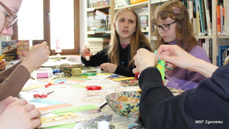 Zbliżenie na dłonie uczestników pracujących przy stole z&nbsp;materiałami plastycznymi. Widoczne są kolorowe paski papieru, wstążki, klej oraz gotowe i&nbsp;tworzone zakładki do książek. Zdjęcie pokazuje fragment spotkania Młodzieżowego Klubu „Książkowi Wędrowcy”, poświęconego promocji czytelnictwa i&nbsp;Światowemu Dniu Książki i&nbsp;Praw Autorskich.