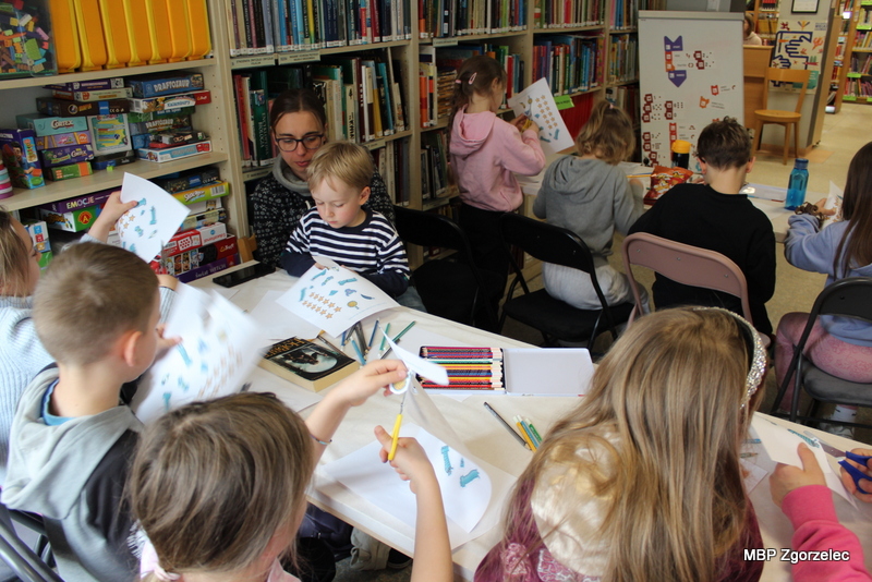 Na zdjęciu widoczna jest grupa dzieci pracujących przy stole w&nbsp;bibliotece. Uczestnicy trzymają kartki z&nbsp;wydrukowanymi elementami, które wycinają i&nbsp;doklejają do rysunków statku kosmicznego Scottie Go. Zajęcia rozwijają kreatywność i&nbsp;sprawność manualną.