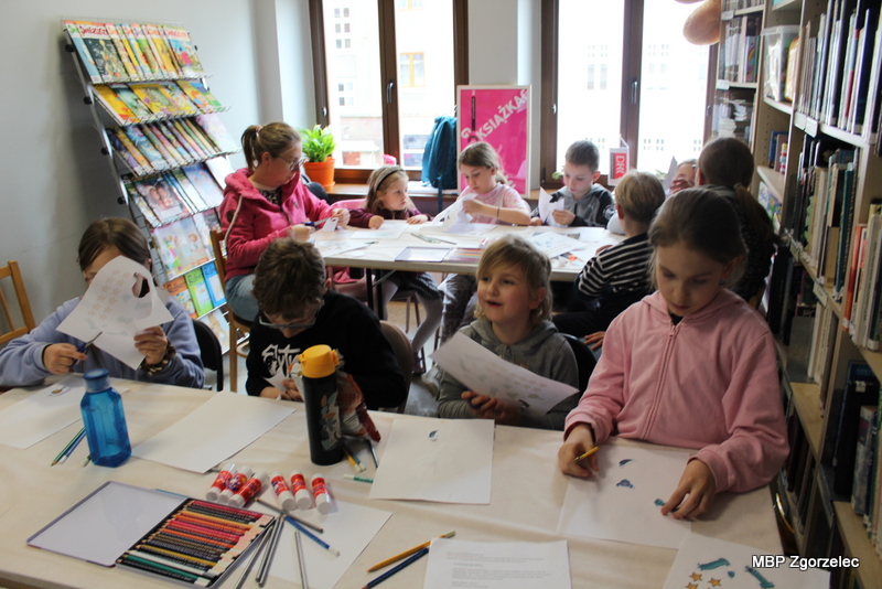 Zdjęcie przedstawia dzieci siedzące przy stołach ustawionych przy oknach biblioteki. Na blatach znajdują się kartki, kredki i&nbsp;kleje. Uczestnicy spotkania wykonują prace plastyczne inspirowane kosmiczną tematyką spotkania Dyskusyjnego Klubu Książki.