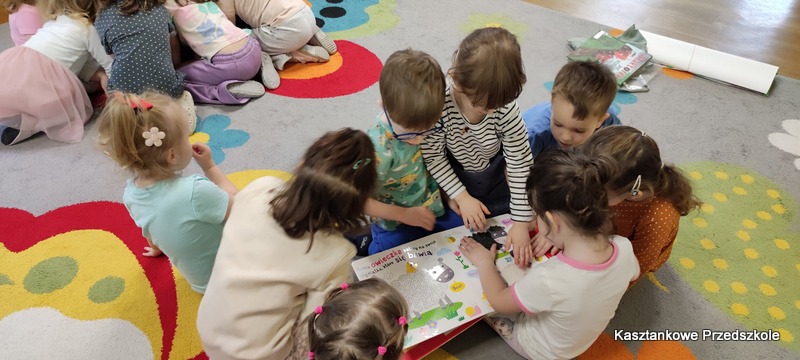 Grupa dzieci siedząca na dywanie przegląda interaktywną książkę o&nbsp;zwierzętach wiejskich, poznając ilustracje i&nbsp;elementy dotykowe.