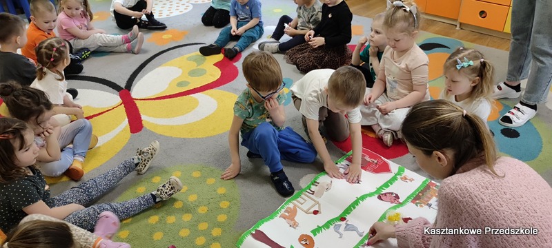 Dzieci wykonują wspólną pracę plastyczną na dywanie, przedstawiającą zwierzęta, krajobraz i&nbsp;zabudowania wiejskie, w&nbsp;ramach zajęć tematycznych.