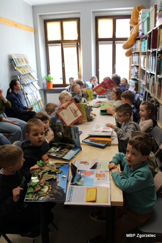 Grupa dzieci siedzi przy długim stole w&nbsp;czytelni biblioteki i&nbsp;przegląda interaktywne książki z&nbsp;dużymi ilustracjami. Na stole leżą otwarte publikacje, a&nbsp;w tle znajdują się regały z&nbsp;księgozbiorem oraz okna.