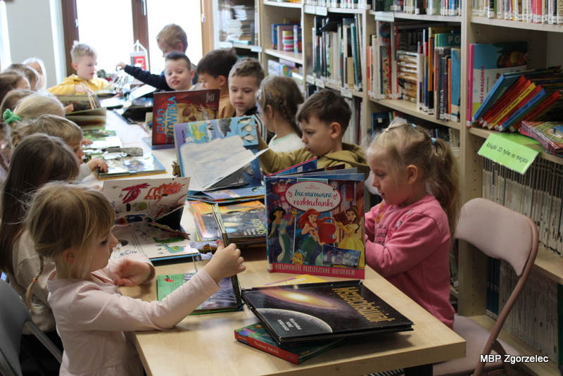 Grupa dzieci z&nbsp;przedszkola siedzi przy długim stole w&nbsp;czytelni biblioteki i&nbsp;przegląda kolorowe książki dla najmłodszych, w&nbsp;tym książki interaktywne i&nbsp;trójwymiarowe. W&nbsp;tle widoczne regały z&nbsp;księgozbiorem.