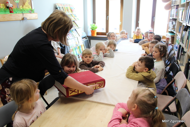 Prowadząca zajęcia prezentuje dzieciom dużą książkę podczas lekcji bibliotecznej. Dzieci siedzą przy stole i&nbsp;słuchają prezentacji w&nbsp;przestrzeni czytelni biblioteki.