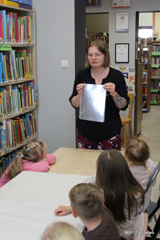 Bibliotekarka pokazuje dzieciom kartkę zapisaną alfabetem Braille’a, omawiając książki przeznaczone dla osób niewidomych. Dzieci siedzą przy stole w&nbsp;czytelni biblioteki.