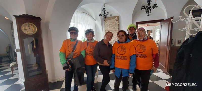 Grupa uczestników rajdu rowerowego w&nbsp;pomarańczowych koszulkach stoi we wnętrzu zabytkowego budynku; widoczne sklepienia, zegar stojący i&nbsp;elementy wystroju.
