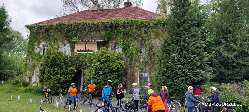 Grupa uczestników rajdu rowerowego z&nbsp;rowerami zatrzymała się przed zabytkowym budynkiem porośniętym bluszczem (Dwór w&nbsp;Pokrzywniku); obiekt otoczony jest zielenią.