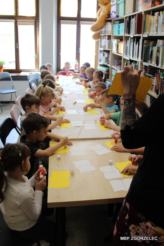 Przedszkolaki siedzą przy długim stole w&nbsp;bibliotece i&nbsp;wykonują prace plastyczne, przyklejając papierowe elementy. Na stole leżą kartki, kleje i&nbsp;materiały do zajęć, w&nbsp;tle regały z&nbsp;książkami i&nbsp;okna.