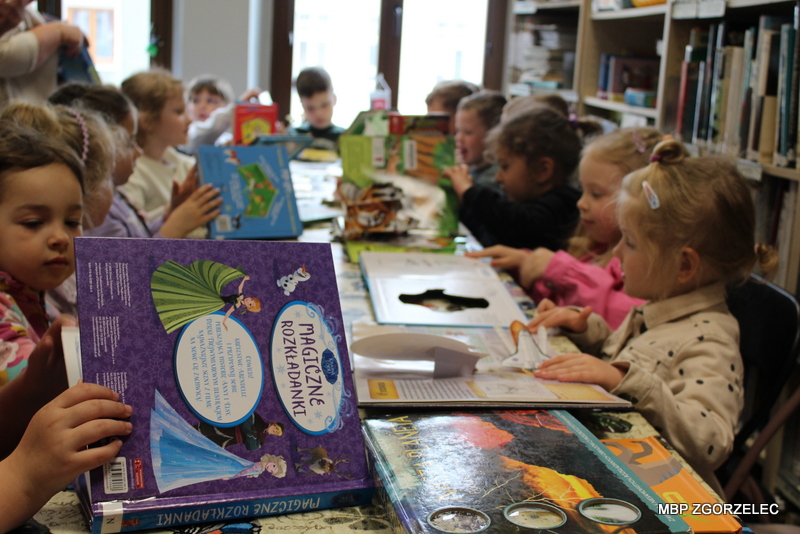 Dzieci siedzą przy stole w&nbsp;bibliotece i&nbsp;wspólnie oglądają książki dla najmłodszych, w&nbsp;tym książki trójwymiarowe pop‑up. W&nbsp;tle regały z&nbsp;księgozbiorem.