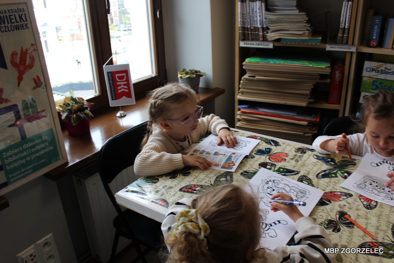 Przedszkolaki siedzą przy stole w bibliotece i kolorują obrazki przedstawiające zwierzęta. Na stole leżą kredki i kartki, w tle widoczne są regały z książkami. Przedszkolaki siedzą przy stole w bibliotece i kolorują obrazki przedstawiające zwierzęta. Na stole leżą kredki i kartki, w tle widoczne są regały z książkami.