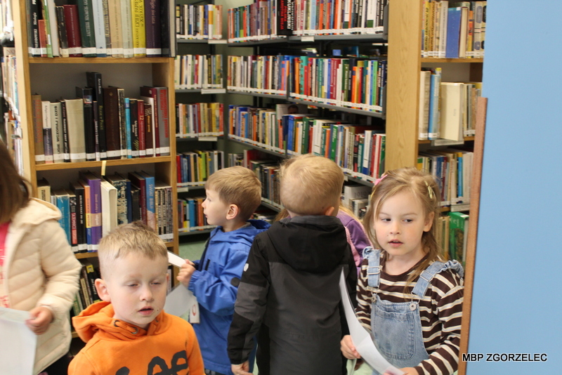 Przedszkolaki stoją między regałami w bibliotece i oglądają książki na półkach. Dzieci trzymają kartki i rozglądają się po księgozbiorze. Przedszkolaki stoją między regałami w bibliotece i oglądają książki na półkach. Dzieci trzymają kartki i rozglądają się po księgozbiorze.