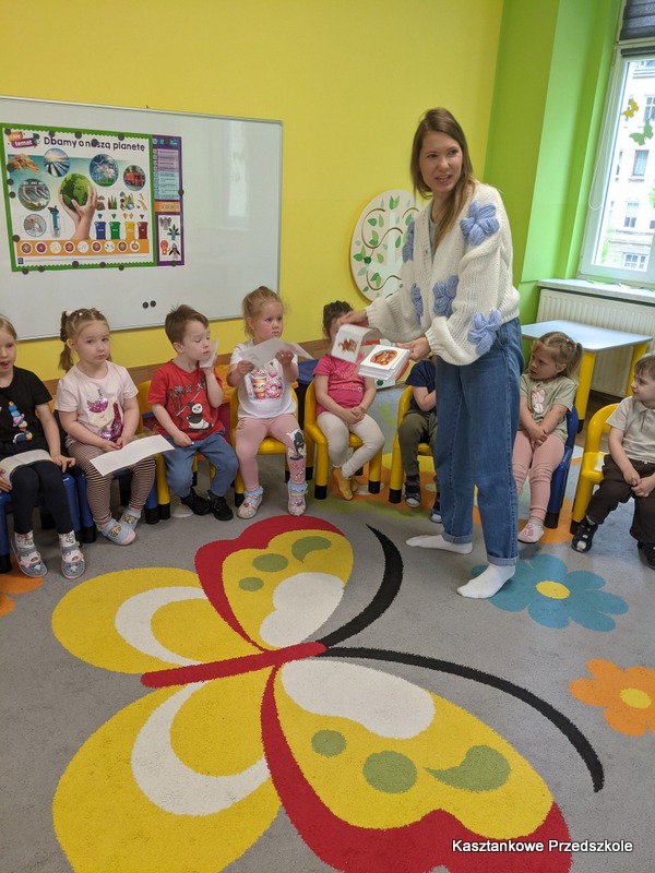 Dzieci siedzą na krzesełkach w&nbsp;sali przedszkolnej, a&nbsp;bibliotekarka prezentuje książkę podczas głośnego czytania. Na ścianach znajdują się plansze edukacyjne, a&nbsp;na podłodze kolorowy dywan.