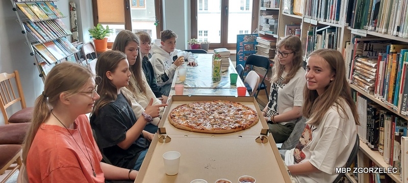 Grupa uczestników spotkania Młodzieżowego Klubu Książki siedzi przy długim stole w bibliotece. Na stole znajduje się duża pizza oraz kubki z napojami. Wokół widoczne są półki z książkami, charakterystyczne dla przestrzeni bibliotecznej. Grupa uczestników spotkania Młodzieżowego Klubu Książki siedzi przy długim stole w bibliotece. Na stole znajduje się duża pizza oraz kubki z napojami. Wokół widoczne są półki z książkami, charakterystyczne dla przestrzeni bibliotecznej.