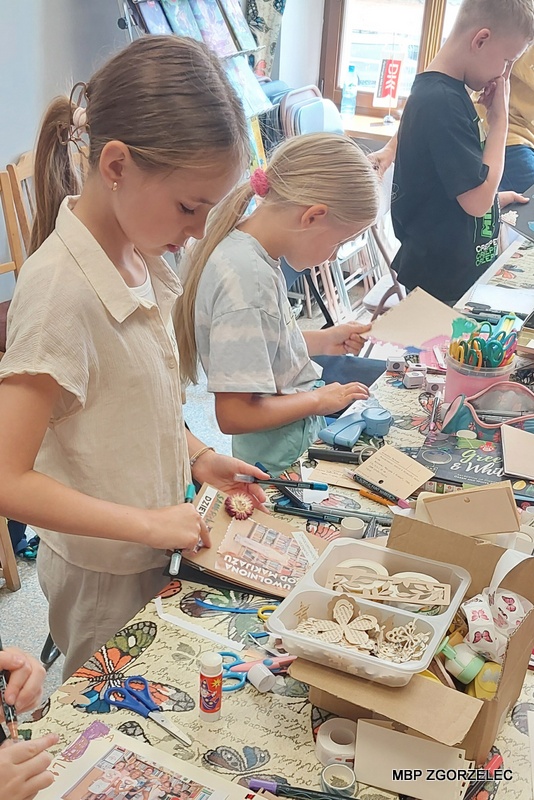Uczestnicy warsztatów scrapbookingowych stoją przy stole w bibliotece i wycinają oraz dekorują elementy do swoich albumów „Księga moich marzeń”; na stole widoczne są nożyczki, kleje, taśmy ozdobne oraz wycinki z czasopism i książek przygotowane do pracy twórczej. Uczestnicy warsztatów scrapbookingowych stoją przy stole w bibliotece i wycinają oraz dekorują elementy do swoich albumów „Księga moich marzeń”; na stole widoczne są nożyczki, kleje, taśmy ozdobne oraz wycinki z czasopism i książek przygotowane do pracy twórczej.