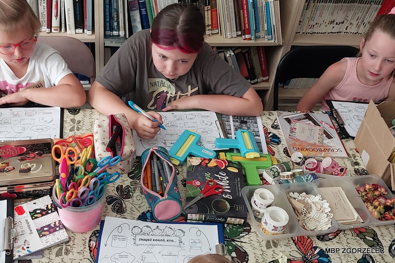 Uczestnicy warsztatów scrapbookingowych siedzą przy stole w bibliotece; na stole widoczne są nożyczki, taśmy ozdobne, stemple oraz wycinki z czasopism i książek. Uczestnicy warsztatów scrapbookingowych siedzą przy stole w bibliotece; na stole widoczne są nożyczki, taśmy ozdobne, stemple oraz wycinki z czasopism i książek.