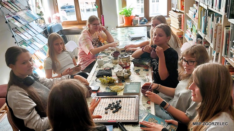 Uczestnicy Młodzieżowego Klubu „Książkowi Wędrowcy” przy stole z&nbsp;książkami, napojami i&nbsp;planszą do Scrabble podczas spotkania w&nbsp;bibliotece.