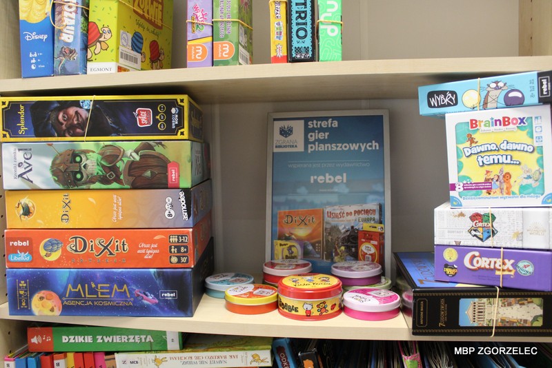 Na regale w&nbsp;Miejskiej Bibliotece Publicznej w&nbsp;Zgorzelcu prezentowana jest kolekcja gier planszowych udostępnionych w&nbsp;ramach programu „Zgrane Biblioteki”. Widoczne są pudełka gier, m.in. „Splendor”, „Avel”, „Dixit”, „MLEM: Agencja Kosmiczna”, „Trio”, „Wydry” i&nbsp;„Cortex”. Pośrodku znajduje się tabliczka z&nbsp;napisem „Strefa gier planszowych”, a&nbsp;obok ustawiono puszki z&nbsp;grą „Dobble”. Kolekcja gier jest wyeksponowana na bibliotecznym regale wśród książek.
